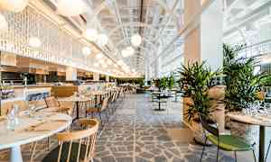 En modern och ljus restaurang med bord och stolar samt gröna växter. Takhöjd och belysning bidrar till en luftig atmosfär. (AI-genererad text)