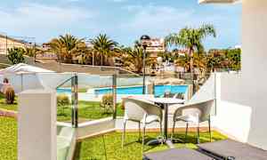 Utsikt från en terrass med solstolar, bord och stolar mot en bassäng och omgivande grönska. (AI-genererad text)