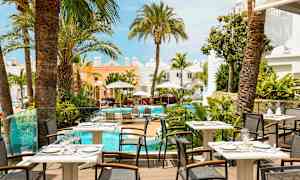 Bild av en restaurangterrass med utsikt över en pool omgiven av palmer och byggnader i ljusa färger. (AI-genererad text)