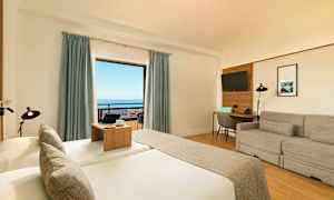 Ett hotellrum med en säng, soffa, skrivbord och ett stort fönster med utsikt över havet. (AI-genererad text)