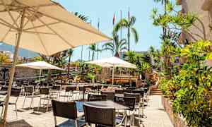 Bild av en uteplats med bord, stolar och parasoller i en tropisk miljö. (AI-genererad text)