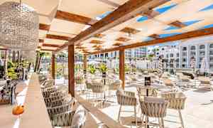 En utomhusbar med stolar och bord under en pergola, omgiven av solstolar och grönska. (AI-genererad text)