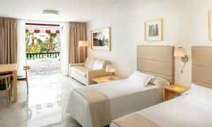 Hotellrum med två enkelsängar, en soffa, ett bord med stolar och ett fönster mot en balkong. (AI-genererad text)