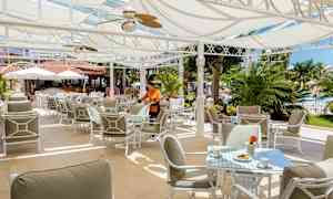 Utomhusrestaurang med solskydd, bord och stolar, samt grönska i bakgrunden. (AI-genererad text)