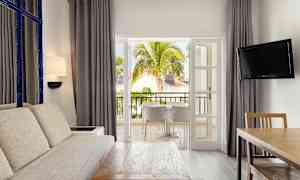 Ett vardagsrum med stor soffa, träbord och en TV, öppnar upp mot en balkong med utsikt över tropiska träd och byggnader. (AI-genererad text)