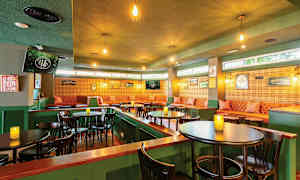 Ett mysigt inrett kafé eller pub med gröna väggar, upplyst av hängande lampor, bruna bord och stolar, samt en sittgrupp med stoppade soffor och väggdekorationer. (AI-genererad text)