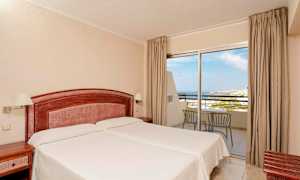 Ett hotellrum med två enkelsängar, ett trähuvudgavel och nattduksbord. Ett glasparti med utsikt mot havet och balkongmöbler. (AI-genererad text)