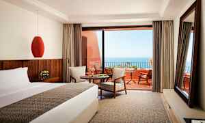 En hotellrum med säng, bord och sittgrupp, med utsikt mot havet från en terrass. (AI-genererad text)