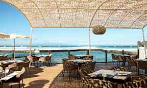 En utomhusrestaurang vid havet med rottingstolar, träbord och en halvt genomskinlig takstruktur. (AI-genererad text)