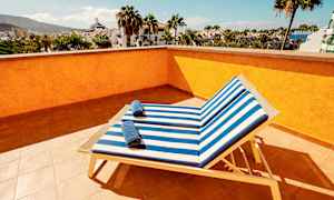 Utsikt från en solterrass med två solstolar med randiga blå och vita dynor samt handdukar på dem, omgivna av palmer och byggnader. (AI-genererad text)