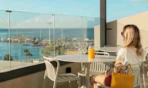 En person sitter vid ett bord med ett glas och en karaff orange juice med utsikt över en småbåtshamn. (AI-genererad text)