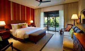 Ett rymligt sovrum med en stor säng, två nattduksbord med lampor, en soffa, en fåtölj och en balkong med utsikt över frodig grönska. (AI-genererad text)