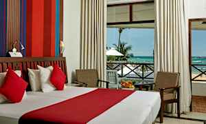 Ett hotellrum med en dubbelsäng med röda och vita sängkläder, en sittgrupp med två stolar och ett bord, och en stor fönsterdörr som visar utsikten mot havet och palmträd. (AI-genererad text)