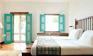 Ett ljust sovrum med en stor säng, vita lakan och turkosa fönsterluckor som släpper in naturligt ljus. (AI-genererad text)