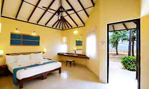 Ett sovrum med högt tak och en fläkt. Rummet har en stor säng med blå dekorativa detaljer, ett skrivbord med en stol samt fönster med ljusa gardiner. Dörren leder ut till en strand med palmer. (AI-genererad text)