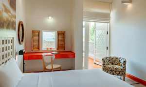 Ett ljust hotellrum med en vit bädd, ett röd bord med en skrivbordsstol och fönsterluckor vid fönstret. Ett mönstrat fåtölj står på andra sidan av rummet. (AI-genererad text)