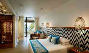 Ett hotellrum med modern inredning, säng med blå och vita textilier, vägg med geometriska mönster, sittgrupp och en balkong med utsikt mot palmer och havet. (AI-genererad text)