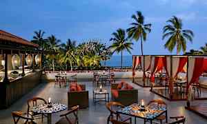 En utomhusrestaurang med bord, stolar och soffor, omgiven av palmer med havet synligt i bakgrunden. (AI-genererad text)