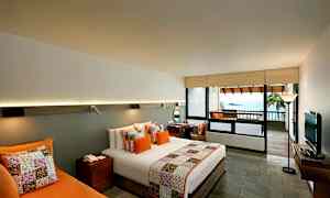 Hotellrum med modern inredning, innehållande en dubbelsäng med färgade kuddar och överkast. Ett skrivbord, en soffa och en TV kompletterar rummet. Det finns en balkong med utsikt över havet. (AI-genererad text)