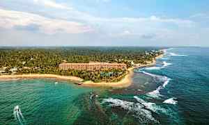 Flygfoto av en tropisk kustlinje med ett hotell nära stranden, omgivet av palmer och hav. (AI-genererad text)