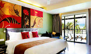 Hotellets rum med en stor säng, färgglada kuddar, en sittgrupp och en balkong med utsikt över en sandstrand omgiven av palmer. (AI-genererad text)