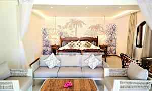 Ett hotellrum med soffa och fåtöljer framför en stor säng med prydnadskuddar och tropisk bakgrund. (AI-genererad text)