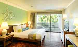 Ett hotellrum med en stor dubbelsäng, träparkettgolv och en balkong med utsikt över palmträd. (AI-genererad text)