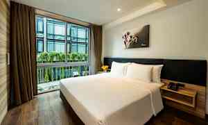 Ett hotellrum med en dubbelsäng, ljust bäddset och trägolv. Stora fönster med utsikt mot en byggnad med växter hängande längs fasaden. (AI-genererad text)