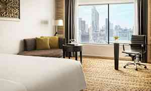 Bild av ett hotellrum med en säng, en soffa, ett skrivbord och en stol. Ett stort fönster visar en vy av en stad. (AI-genererad text)