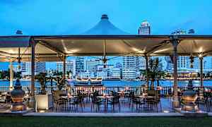 Uteservering med bord och stolar, belägen vid en flod, med en stad i bakgrunden. (AI-genererad text)