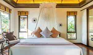 Ett sovrum med en stor säng dekorerad med kuddar och en vit baldakin. Rummet har fönster med utsikt mot grönska. (AI-genererad text)