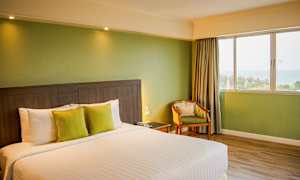 Bild av ett hotellrum med en stor säng, två kuddar, ett nattbord med telefon, en stol, och ett fönster som visar utsikt över havet. (AI-genererad text)