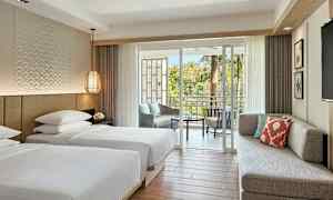 Ett hotellrum med två enkelsängar, en soffa, en balkong med sittmöbler och grönska utanför. Rummet är ljust och moderna möbler. (AI-genererad text)