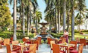 Bilden visar en utomhusrestaurang med bord och stolar, omgivna av palmer. I bakgrunden syns en fontän och en grönskande trädgård. (AI-genererad text)