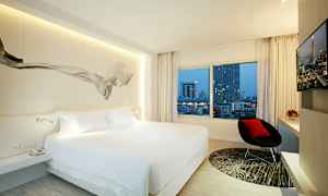 Ett modernt hotellrum med en stor säng, ett fönster med utsikt över stadsbyggnader, en stol och en tv. (AI-genererad text)