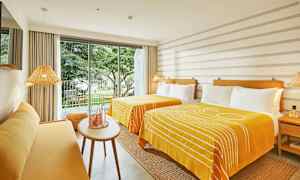 Ett ljust hotellrum med två sängar med gula överkast, en gul soffa och en glasdörr som leder ut till en balkong med utsikt över trädgården. (AI-genererad text)