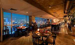 En elegant restaurang med moderna möbler, fönster som visar en stadsutsikt på kvällen och belysning som skapar en mysig atmosfär. (AI-genererad text)