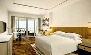 Ett modernt hotellrum med en stor säng, soffa och matbord i ett ljust utrymme. Rummet har en balkong med utsikt och en TV på väggen. (AI-genererad text)