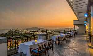 En terrass med uppdukade matbord och stolar, omgiven av en utsikt över en stad vid solnedgången. (AI-genererad text)