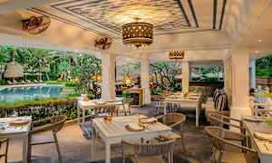 En utomhusrestaurang med vita bord och stolar, omgiven av grönska och med utsikt mot en pool. (AI-genererad text)