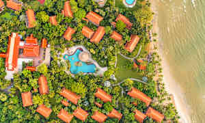 Flygbild över en resort med röda tak omringad av grönska, simbassänger och nära en strand. (AI-genererad text)