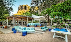 En utomhusterrass med sittgrupper och bord under solskyddstak, omgiven av grönska och med en klippa i bakgrunden. (AI-genererad text)