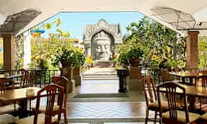 Uteservering med trästolar och bord, omgiven av grönska och dekorativa växter. En arkitektonisk struktur syns i bakgrunden. (AI-genererad text)