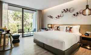 Bild av ett modernt hotellrum med en stor säng, dekorativa kuddar, sittmöbler och ett glasparti som vetter mot en terrass och grönska utanför. (AI-genererad text)