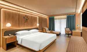 Ett modernt hotellrum med en stor säng, nattduksbord, en fåtölj, ett skrivbord och ett stort fönster med gardiner. (AI-genererad text)