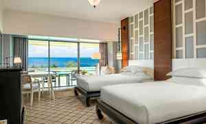 Ett hotellrum med två separata enkelsängar, moderna möbler och en ljus design. Ett stort fönster med utsikt över havet. (AI-genererad text)