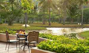 Uteservering med bord och stolar på en träaltan, omgiven av grönska och utsikt över en damm. (AI-genererad text)