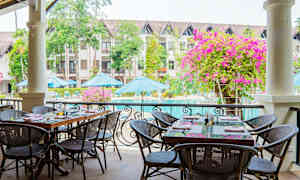 Uteservering med bord och stolar vid en pool. I bakgrunden syns byggnader, parasoller och grönska. (AI-genererad text)