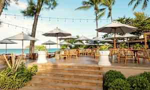 En utomhusrestaurang med bord, stolar och parasoller på en träplattform nära havet. (AI-genererad text)