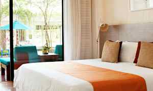 Bild av ett ljust hotellrum med en stor säng klädd i vitt och orange, stora fönster som vetter mot en uteplats med sittgrupp och grönska. (AI-genererad text)